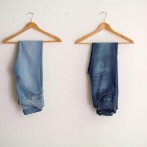 Two pairs of blue denim jeans neatly hanging on wooden hangers against a plain wall.