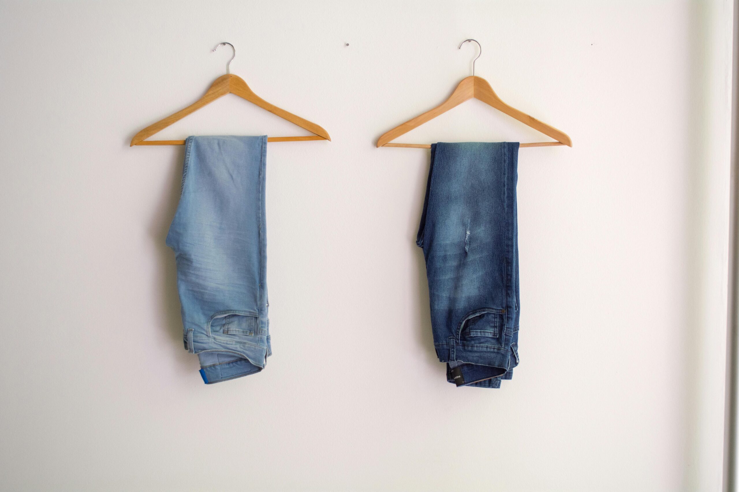 pexels photo 1082528 1082528 Two pairs of blue denim jeans neatly hanging on wooden hangers against a plain wall.